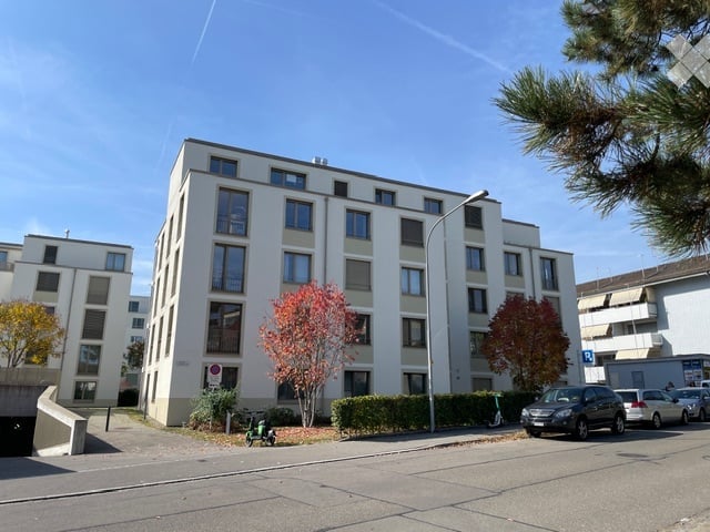 Modern multi-story apartment building with white facade, multiple windows, and surrounding greenery. Several cars are parked in front.