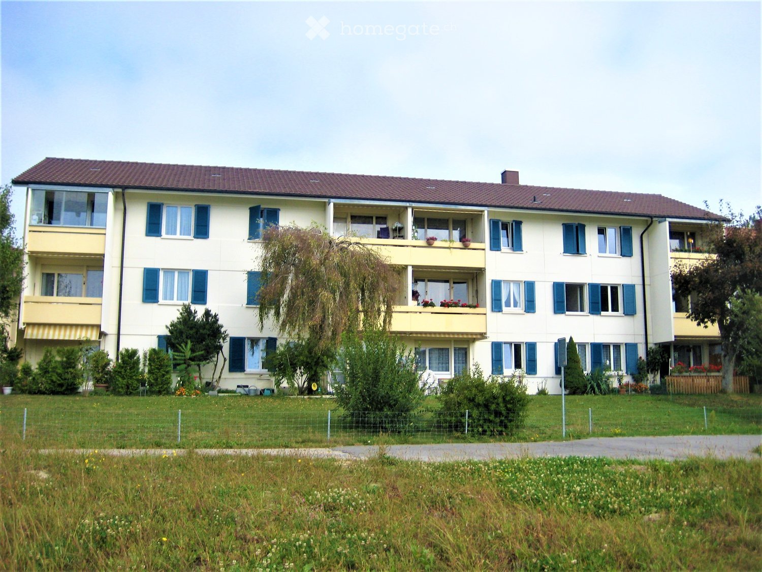 A three-story residential building with light yellow walls, blue shutters, and multiple balconies adorned with potted plants. The building is surrounded by a well-maintained lawn and trees.