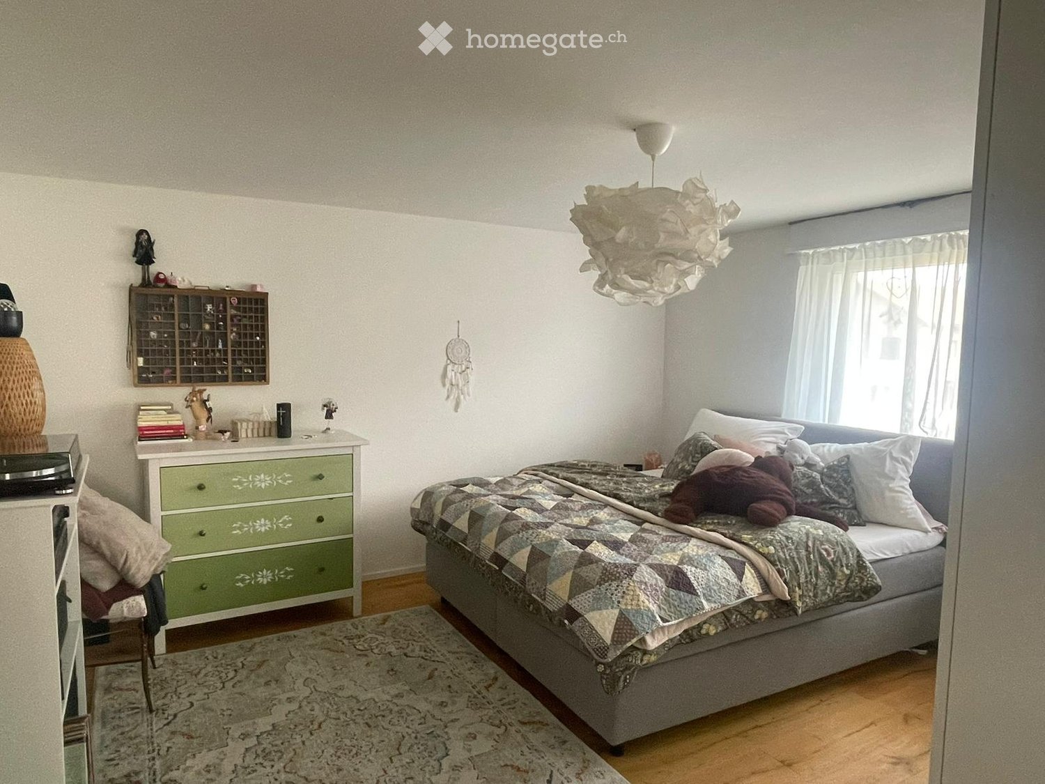 Cozy bedroom with a grey bed, patterned quilt, nightstand, green dresser, and decorative items including a dreamcatcher and storage cabinet.
