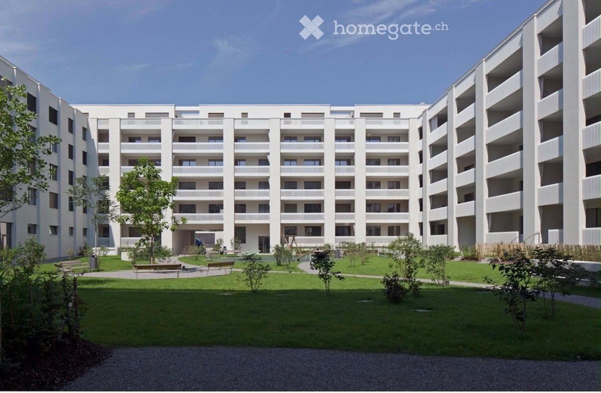 Modern multi-story residential building with white facade, multiple windows, and balconies. The courtyard features green lawns, trees, and benches.