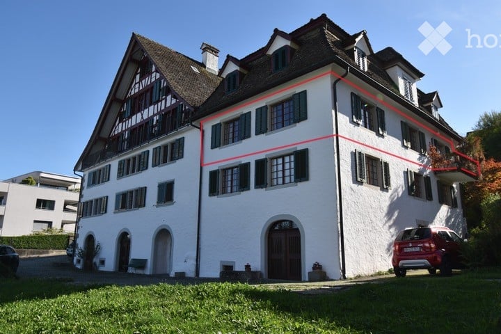 A large, white, multi-story house with a traditional architectural style, featuring multiple windows, a central entrance, and a well-maintained lawn. A red SUV is parked in the driveway.