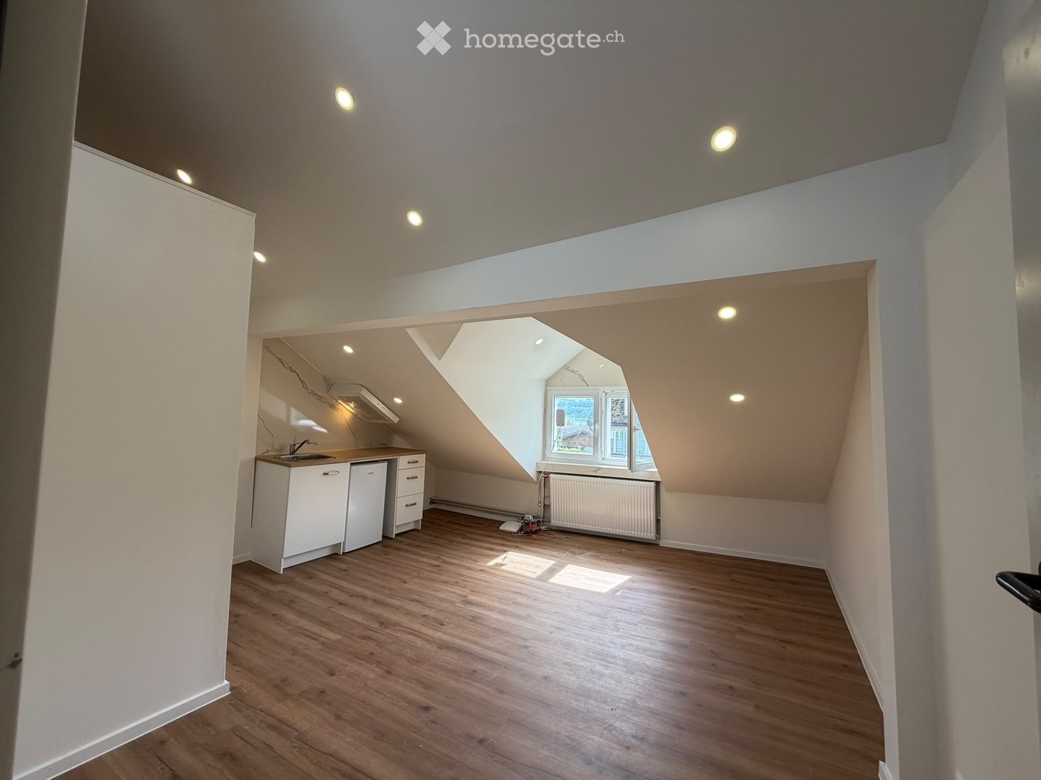 An attic space with wooden flooring, white walls, and a small kitchenette area. The room features skylights and a window, with a radiator and some tools visible.