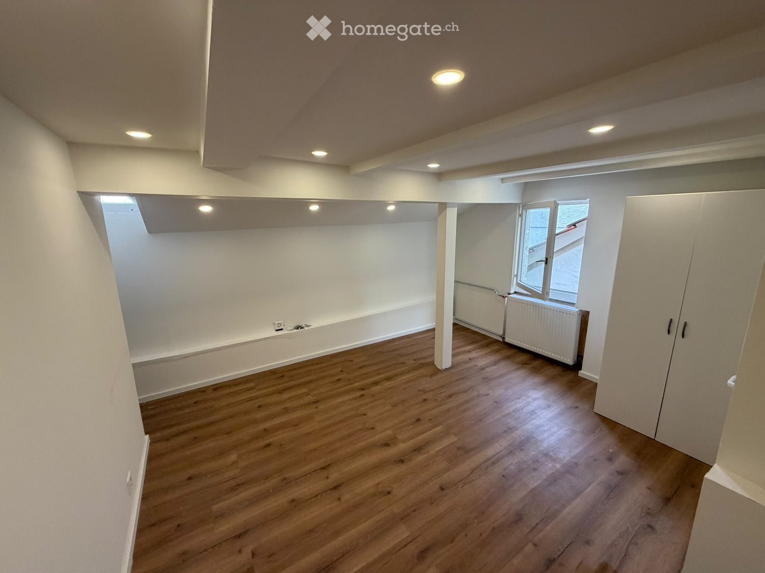 Empty room with wooden flooring, white walls, and recessed lighting. Features a window with blinds and a radiator below it.
