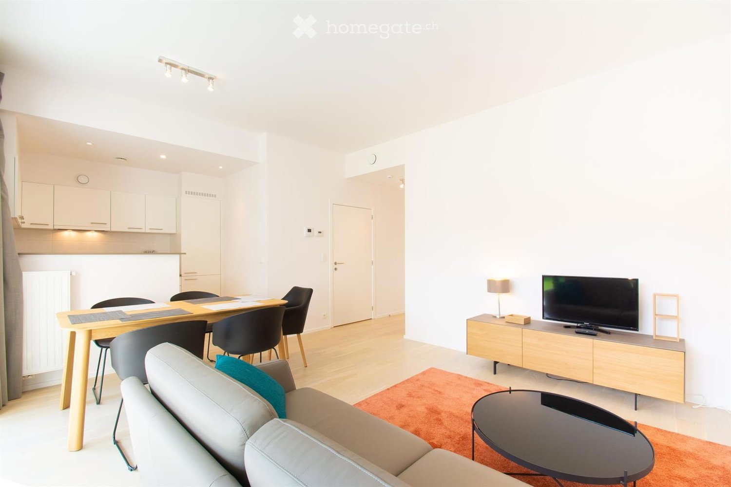 Modern living room with a grey sofa, coffee table, TV stand, dining table, and chairs. The room features a light wooden floor, white walls, and a small kitchen area in the background.