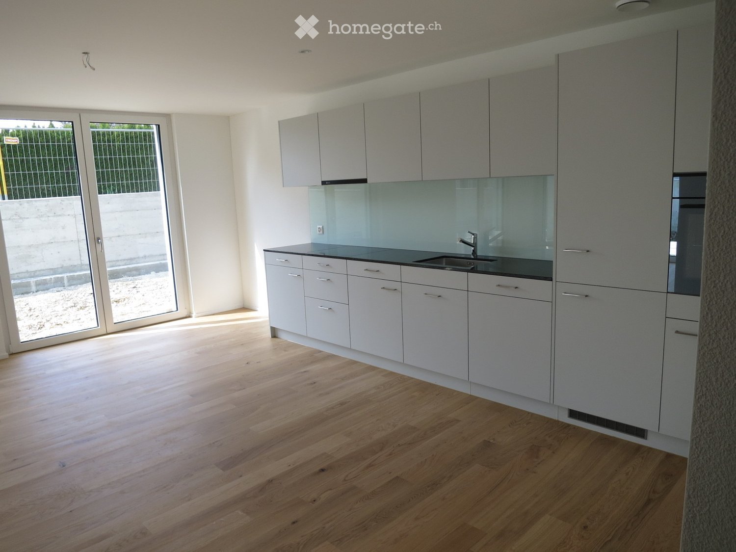Modern kitchen with light grey cabinets, a black countertop, and a sleek sink. The space features light wooden flooring and large glass doors leading outside.