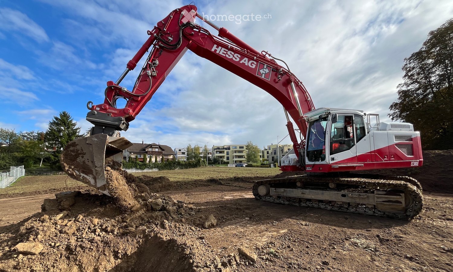 A red excavator is operating on a construction site, moving dirt with its arm extended. The background shows a suburban area with residential buildings and trees.