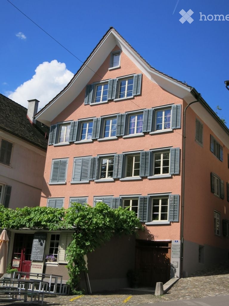 A two-story pink building with multiple windows and green shutters, featuring a triangular roof and a small garden area with a bench and umbrella.