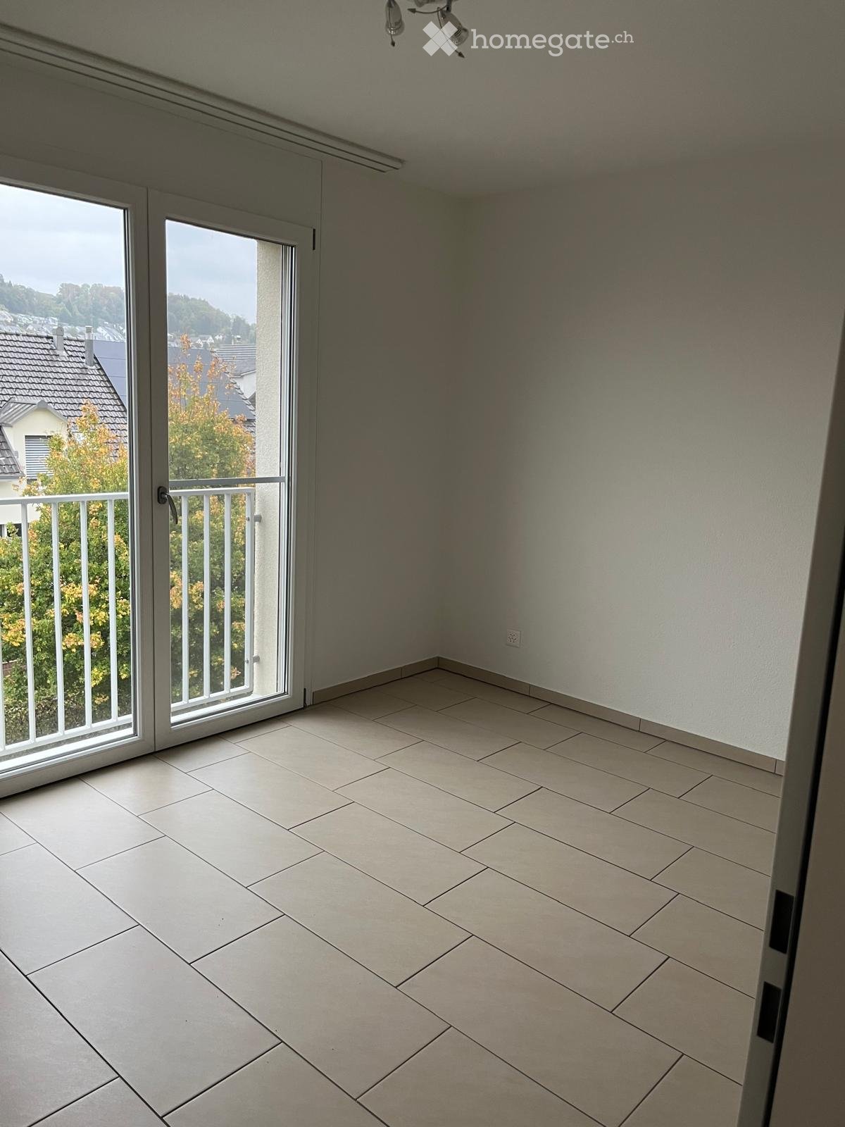 Empty room with tiled floor, white walls, large sliding glass doors leading to a balcony with a metal railing and a view of the neighborhood.