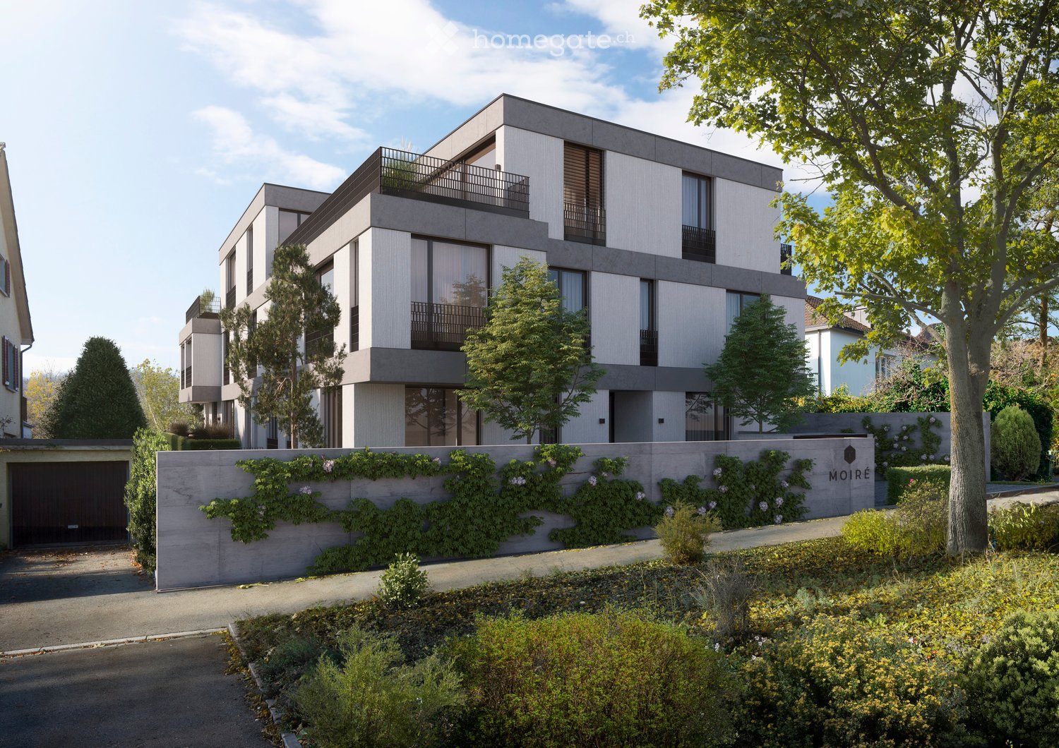 Modern two-story building with balconies, grey facade, and a landscaped garden area in front.