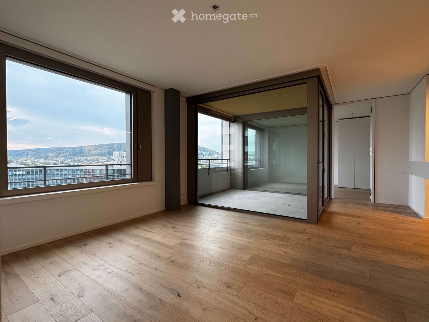 Empty room with wooden flooring, large windows, and a sliding glass door leading to a balcony with a scenic city view.