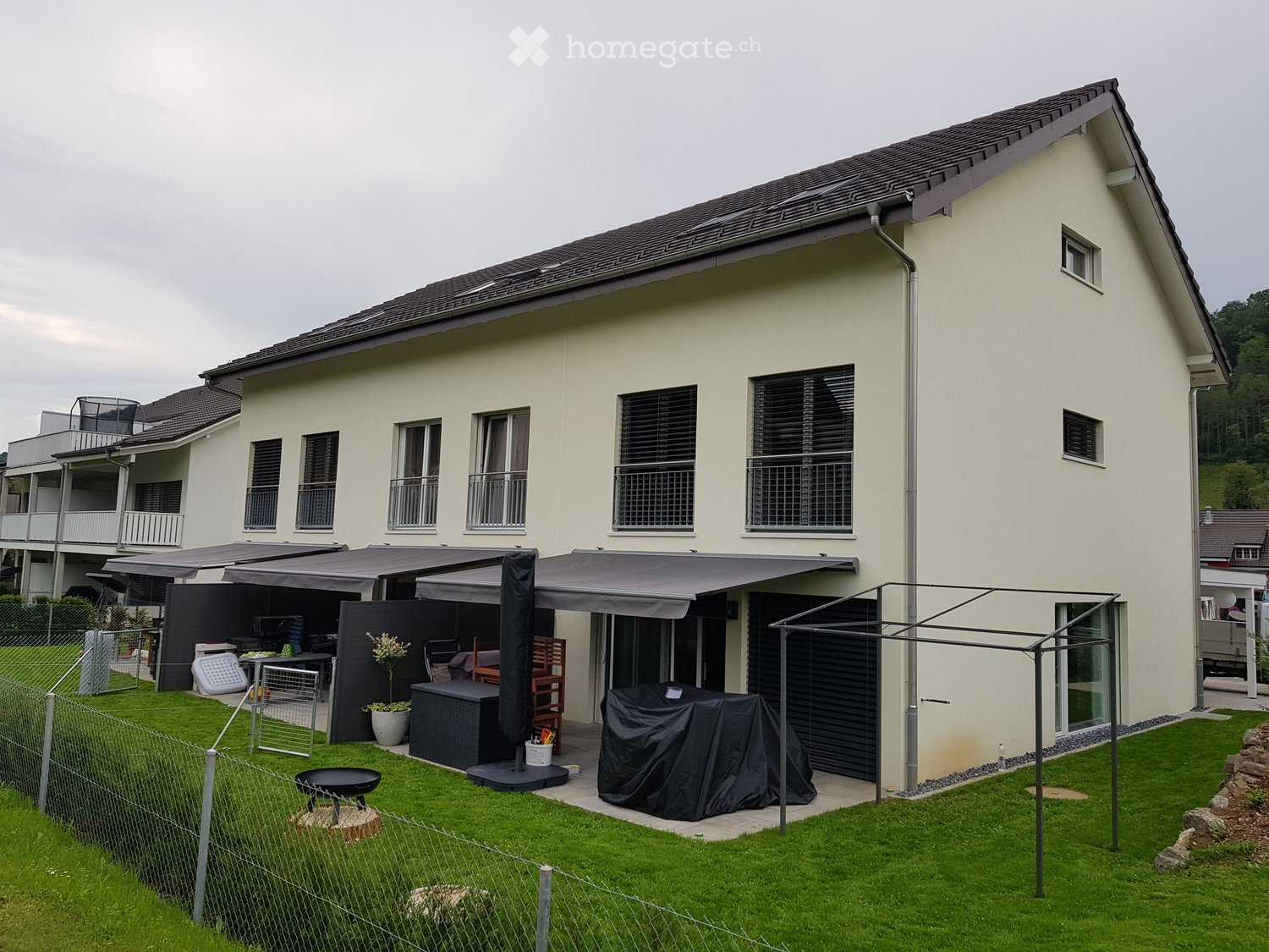 2 story house, beige exterior, black roof, large windows, awnings, garden, outdoor furniture, grill, potted plants
