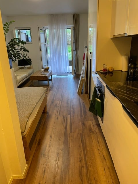 A modern, open-plan living and sleeping area with a kitchen. The space features a wooden floor, a sofa, a coffee table, a bed, and a kitchen with cabinets, an oven, and a fridge. A large window with a view leads to a balcony.