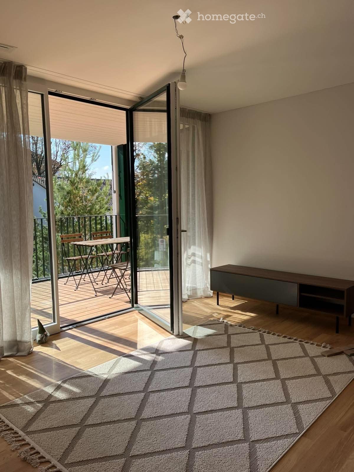 Bright living space with large sliding glass doors opening to a balcony, featuring a wooden floor, a large area rug, a side cabinet, and a small table and chairs on the balcony.