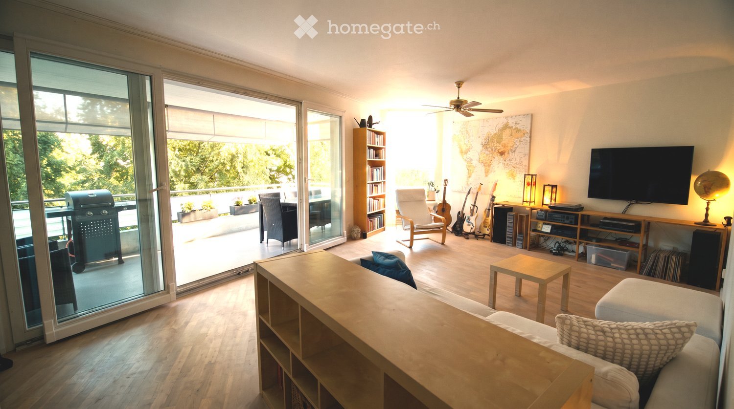 Bright living room with large sliding glass doors leading to a balcony, a white sofa, coffee table, armchair, guitars, bookshelf, and a television on a wooden stand.