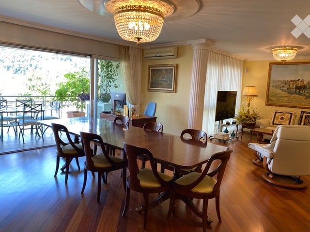 Spacious dining and living area with wooden flooring, large glass doors leading to a balcony, a dining table with chairs, a sofa, a television, and decorative lamps.