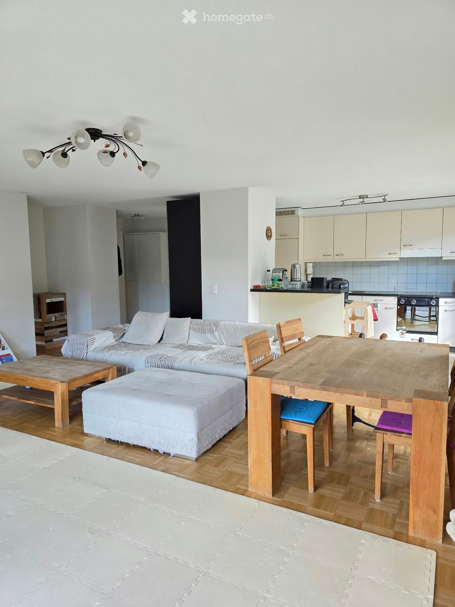 Bright, open-plan living and dining area with wooden flooring, white walls, and modern furniture including a sofa, ottoman, coffee table, dining table with chairs, and a small kitchen area with cabinets and appliances.