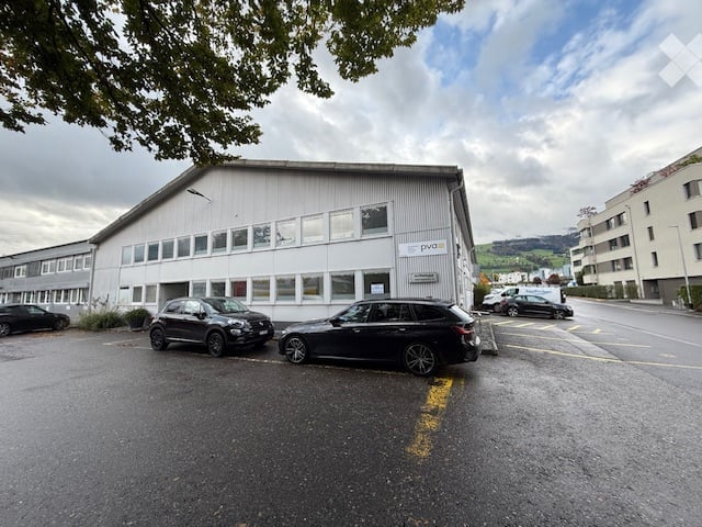 Modern industrial-style building with large windows and a flat roof, surrounded by parked cars in a paved lot.