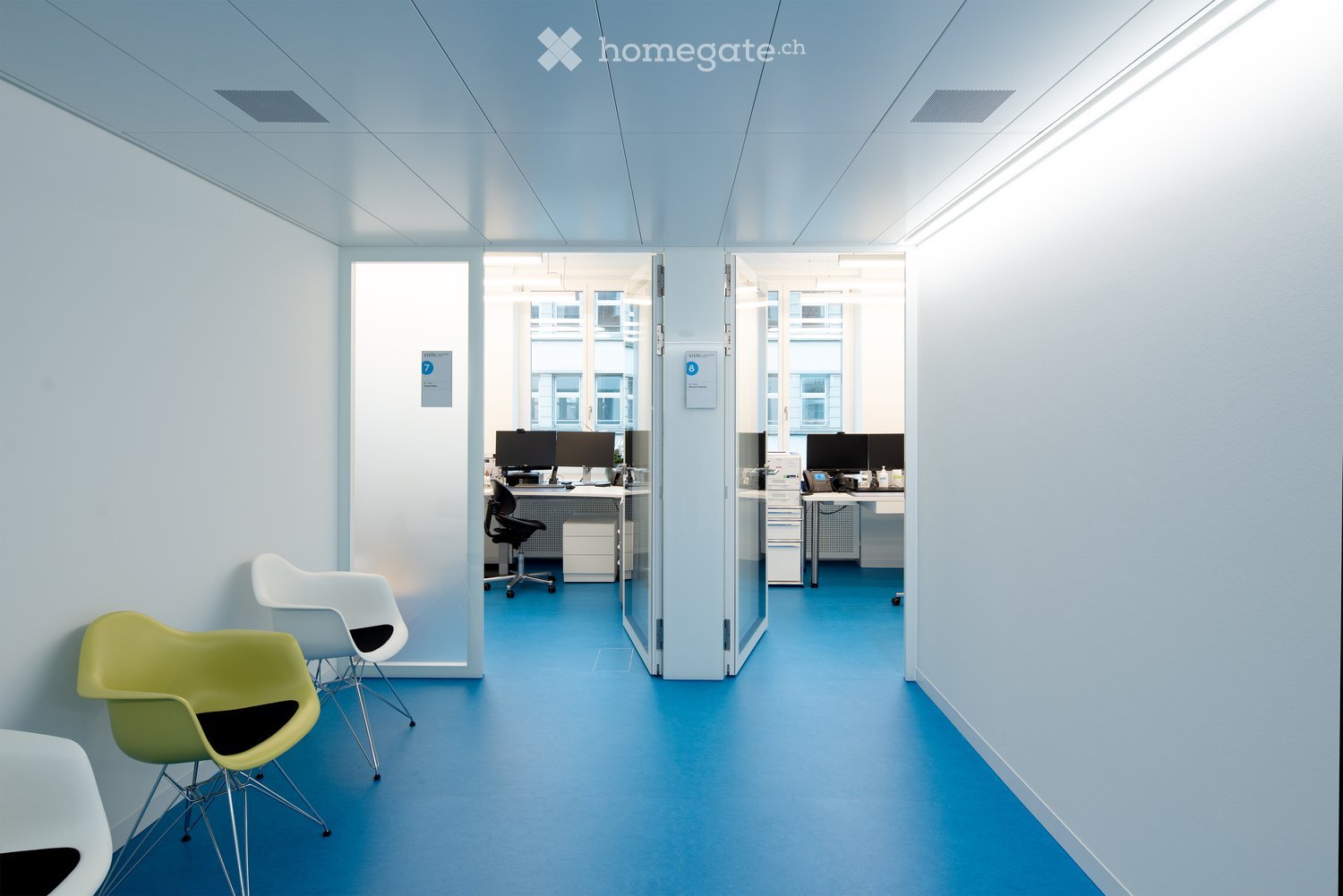 Modern office corridor with light blue walls and floor, featuring white and green chairs, desks with computers, and glass partitions.