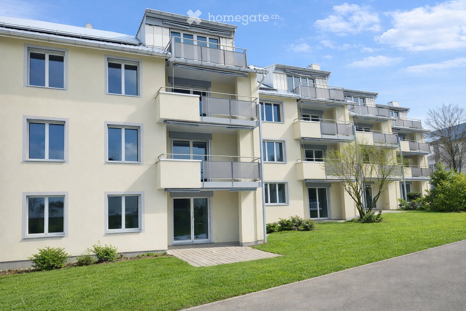 Modern multi-story residential building with light yellow facade, large windows, and multiple balconies. The building is surrounded by a well-maintained lawn and a paved driveway.