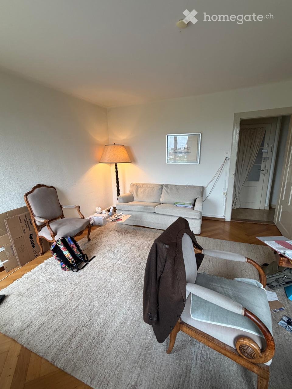 A cozy living room with a light-colored sofa, two armchairs, a coffee table, a floor lamp, and a backpack on the floor. The room features wooden flooring and a large white rug.
