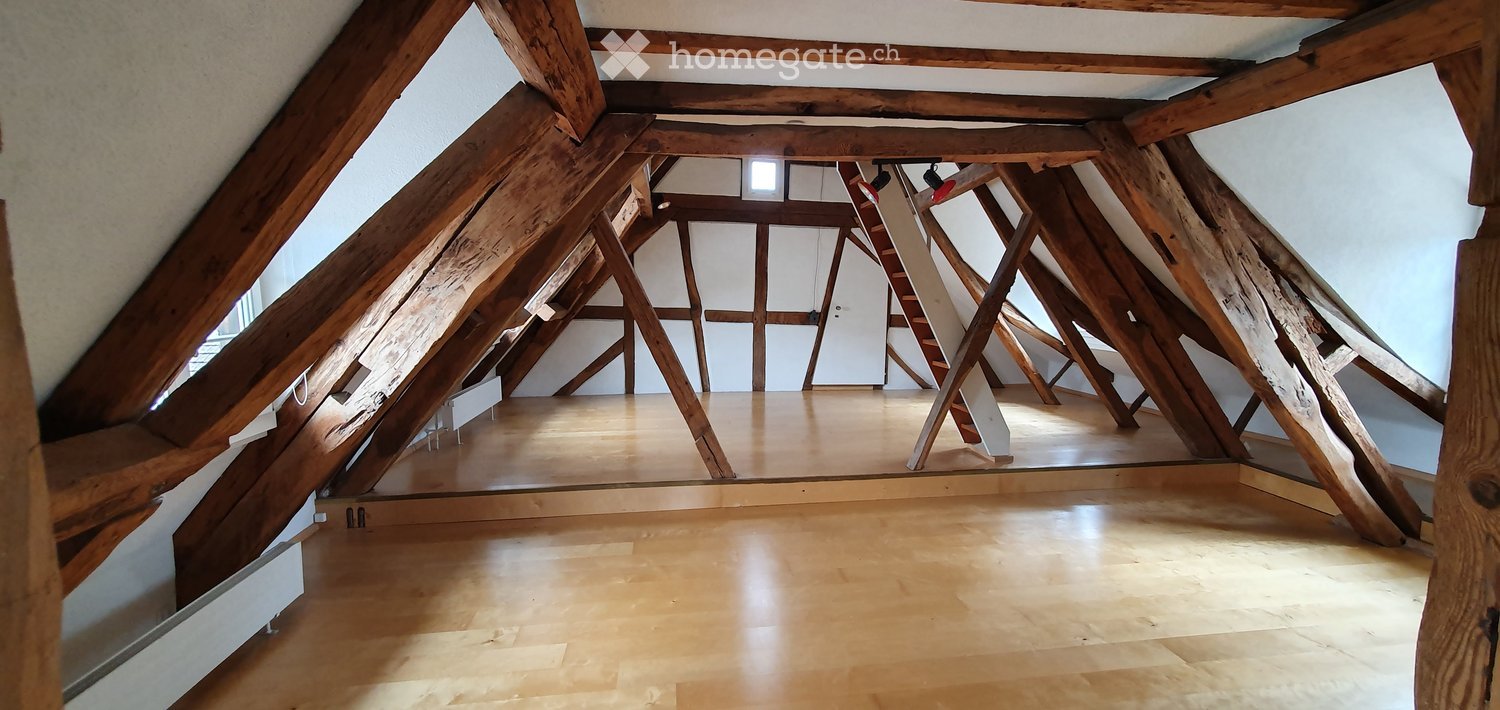An attic space with exposed wooden beams and a wooden floor, featuring large windows and a ladder leading to the upper level.
