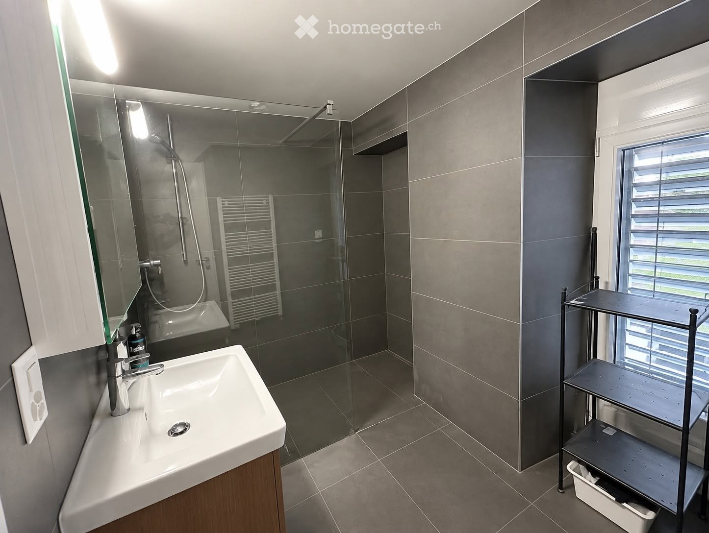 Modern bathroom with a white sink, chrome faucet, large mirror, glass-enclosed shower, and a towel rack. The room features grey tiled walls and a window with blinds.