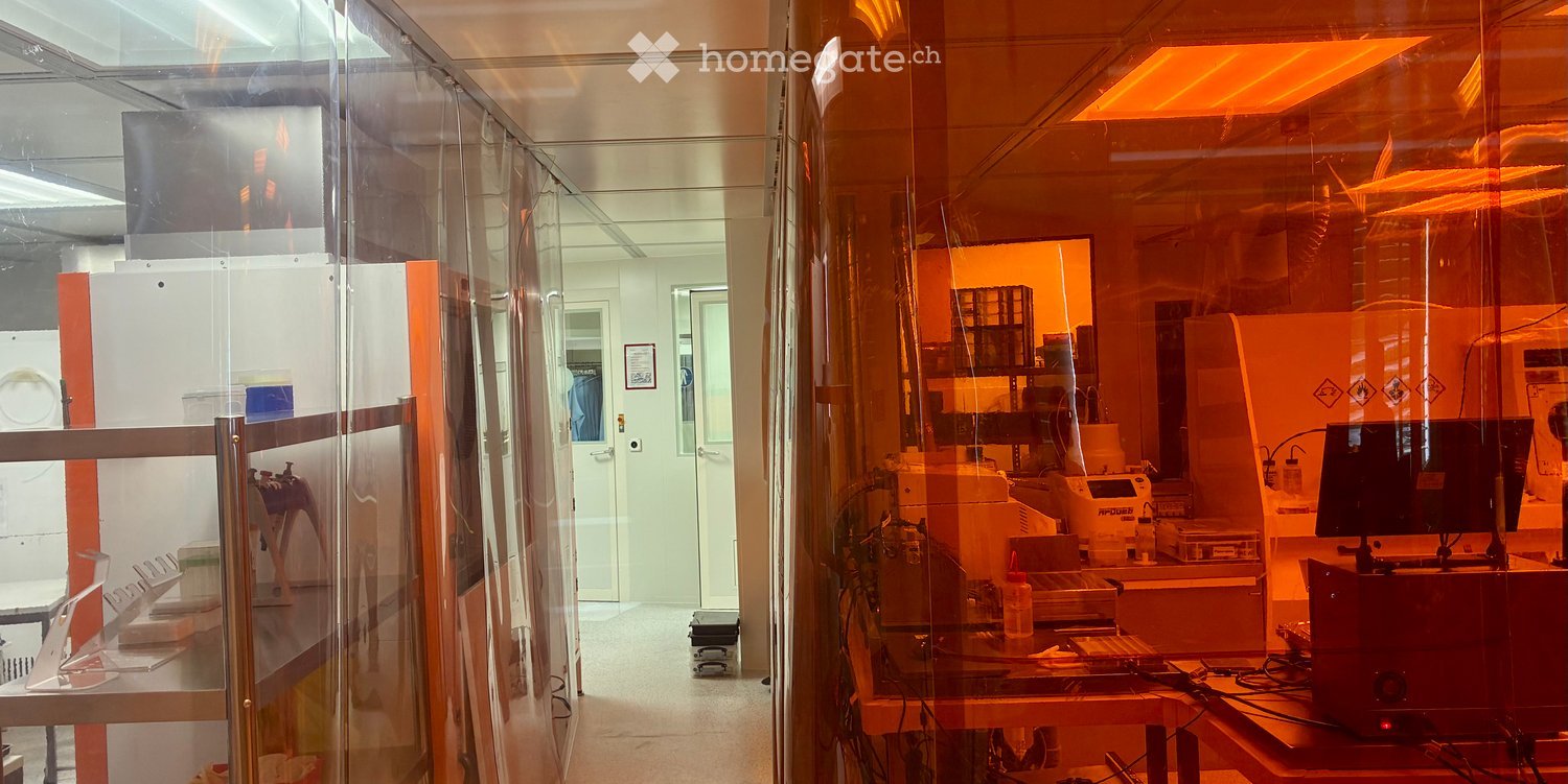 A modern, well-lit room with glass partitions, a table with a screen, a chair, a printer, and a bottle on the table.