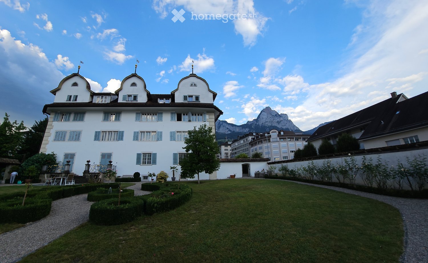 A large, white, multi-story building with classic European architecture, featuring multiple windows, decorative roof elements, and a well-maintained garden with neatly trimmed hedges and pathways.