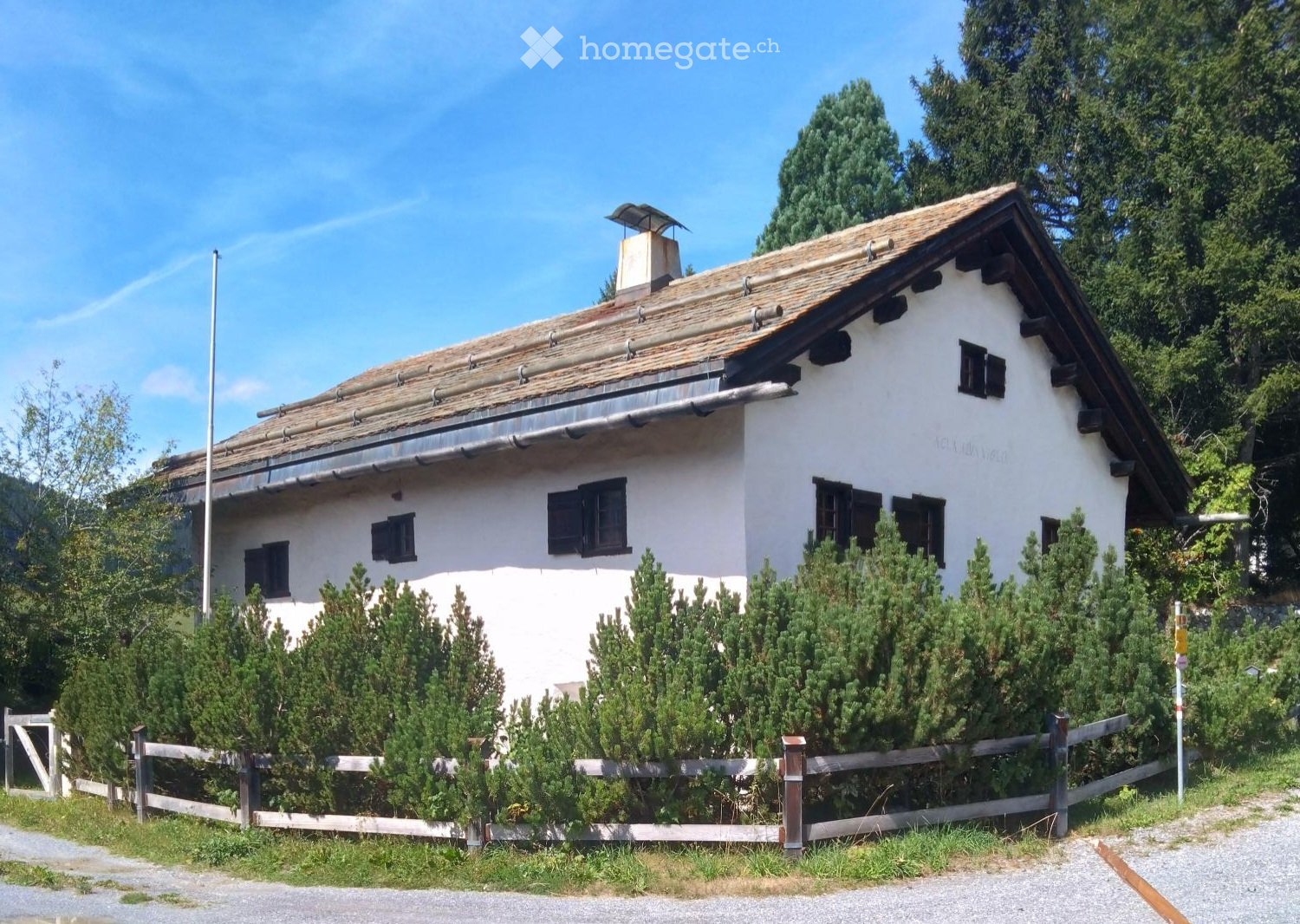 A single-story white house with a tiled roof, surrounded by greenery and a wooden fence.