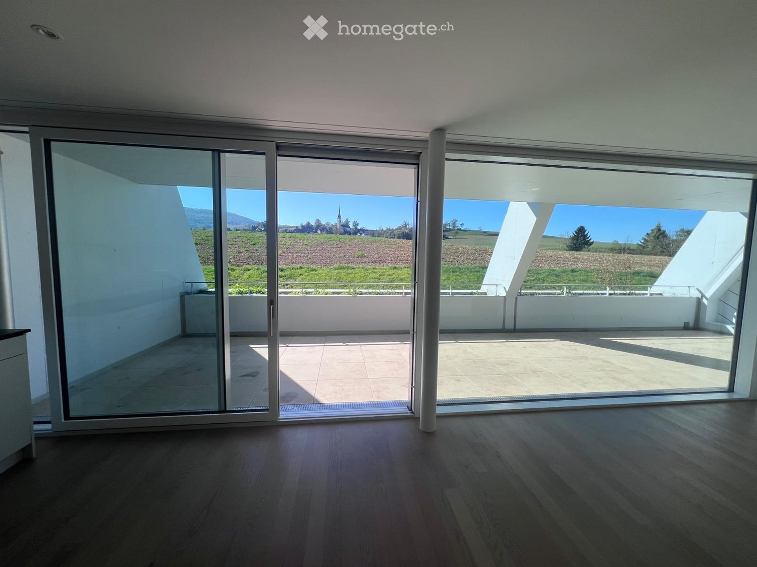 Empty room with wooden flooring, large sliding glass doors leading to a tiled balcony with scenic countryside view.