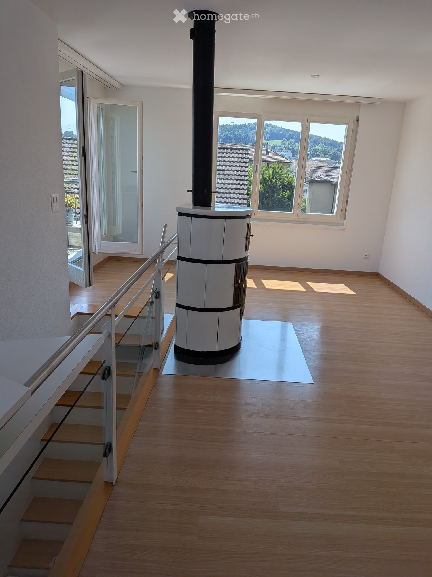 Bright hallway with wooden flooring, a staircase with glass railing, and a central wood-burning stove with a chimney. Large windows offer a scenic view of the surrounding landscape.