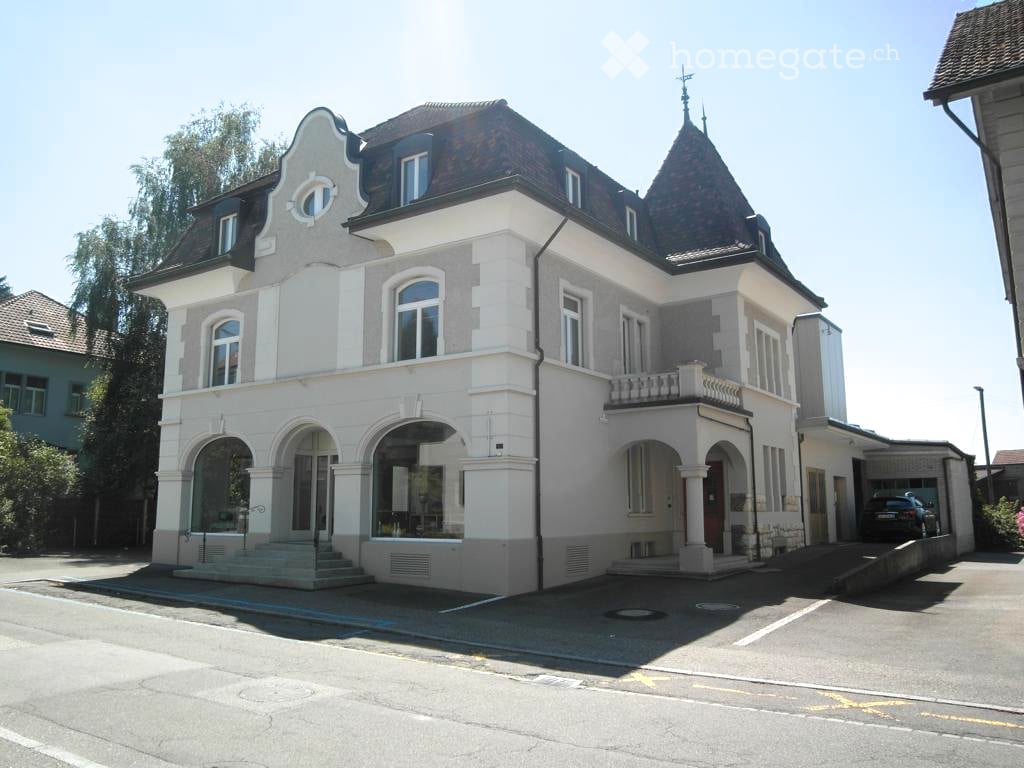 A large, multi-story building with a classic architectural style, featuring arched windows and doors, a prominent balcony, and a garage entrance.