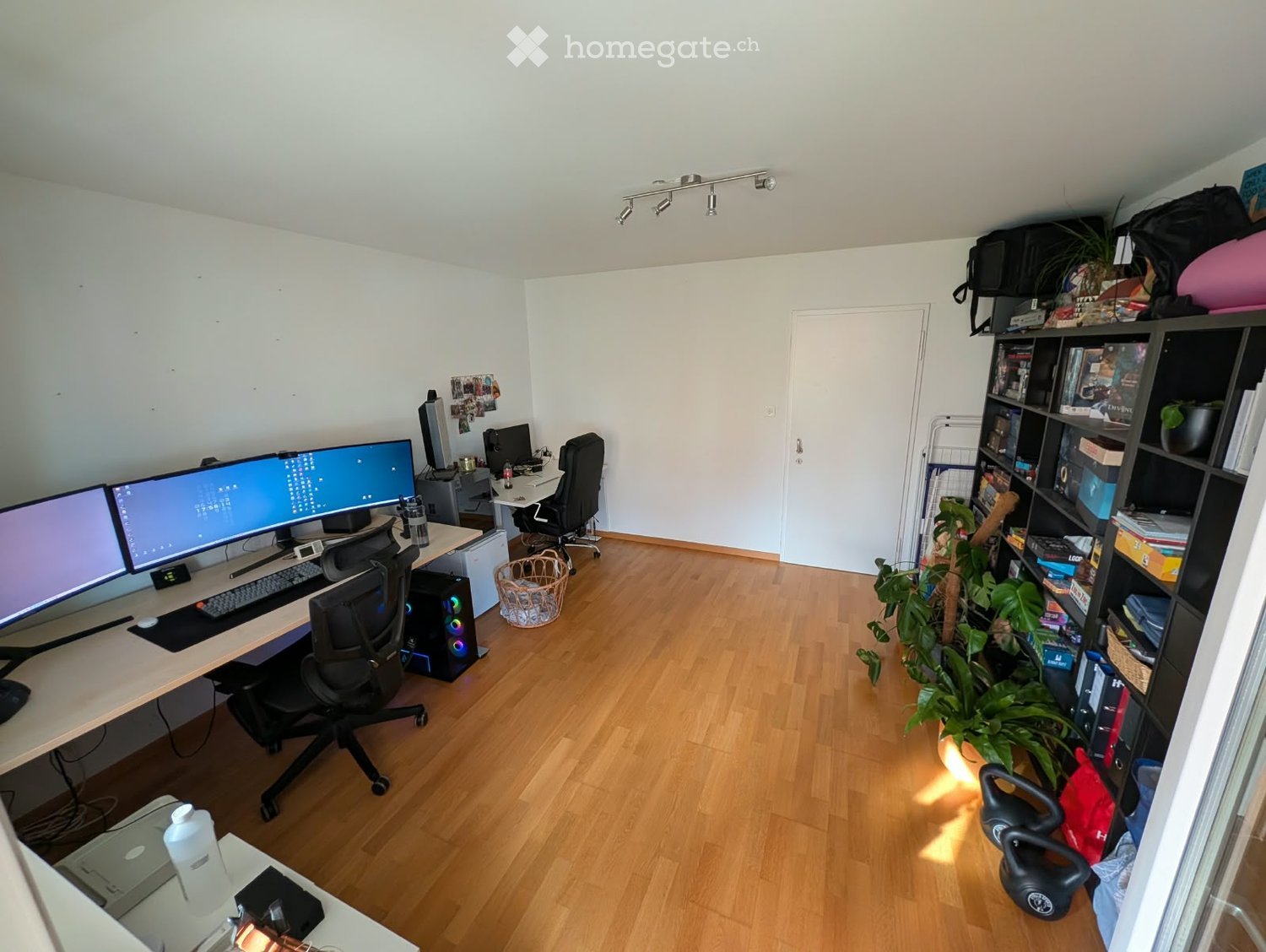 A well-lit room with wooden flooring, a desk setup with dual monitors, a chair, a shelf with books and plants, and a small basket on the floor.