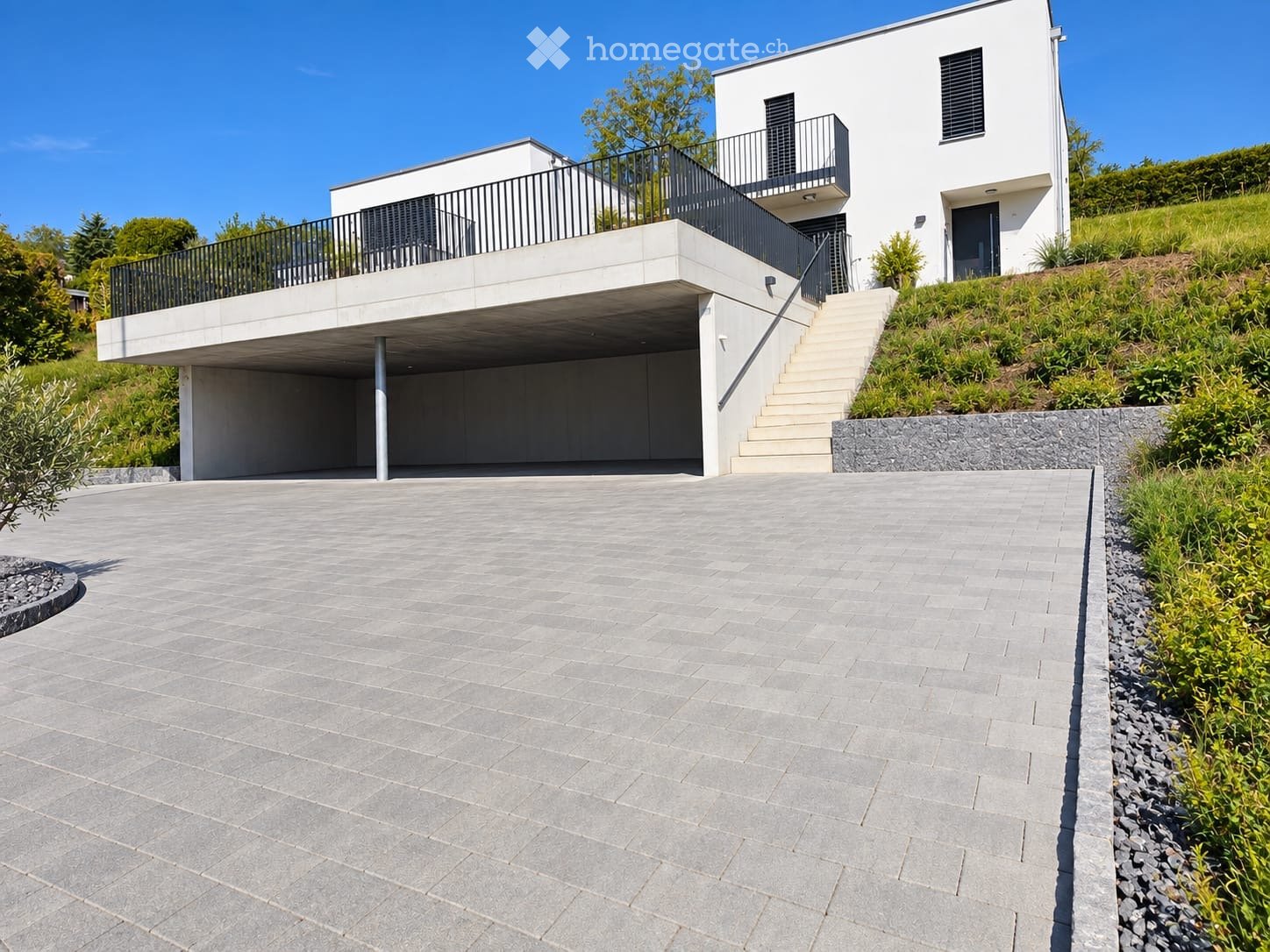 Modern two-story house with white exterior, raised ground floor, garage, balcony, and landscaped garden area.