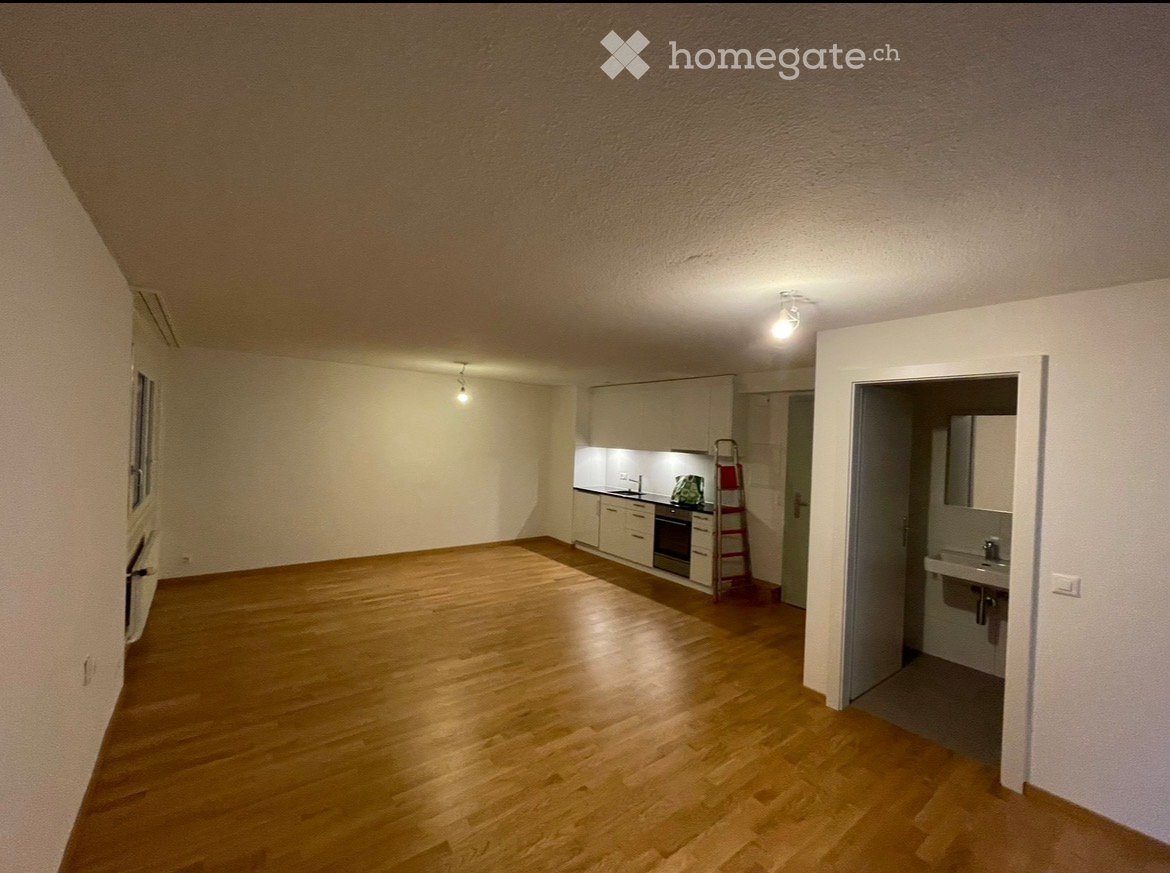 Empty room with wooden flooring, white walls, and a small kitchen area visible in the background. No furniture or personal items are present.