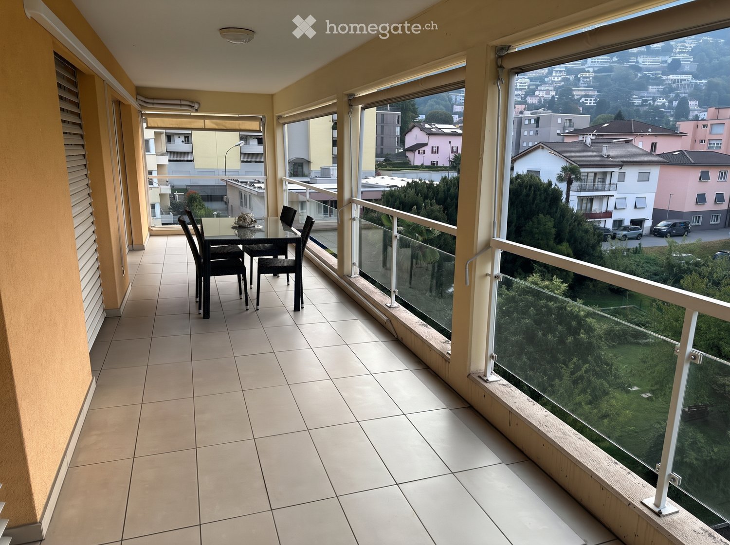 Balcony with tiled floor, glass railing, and a view of the city and greenery. Includes a small table and chairs.