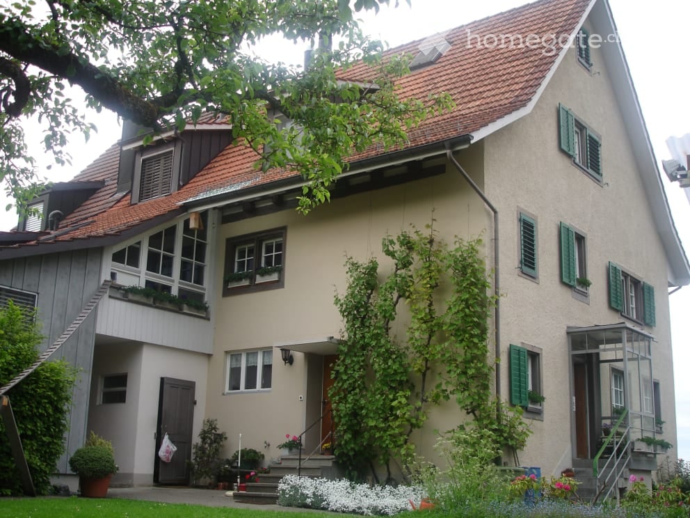 Two-story house with beige walls, green shutters, red-tiled roof, and a small garden with plants and flowers.