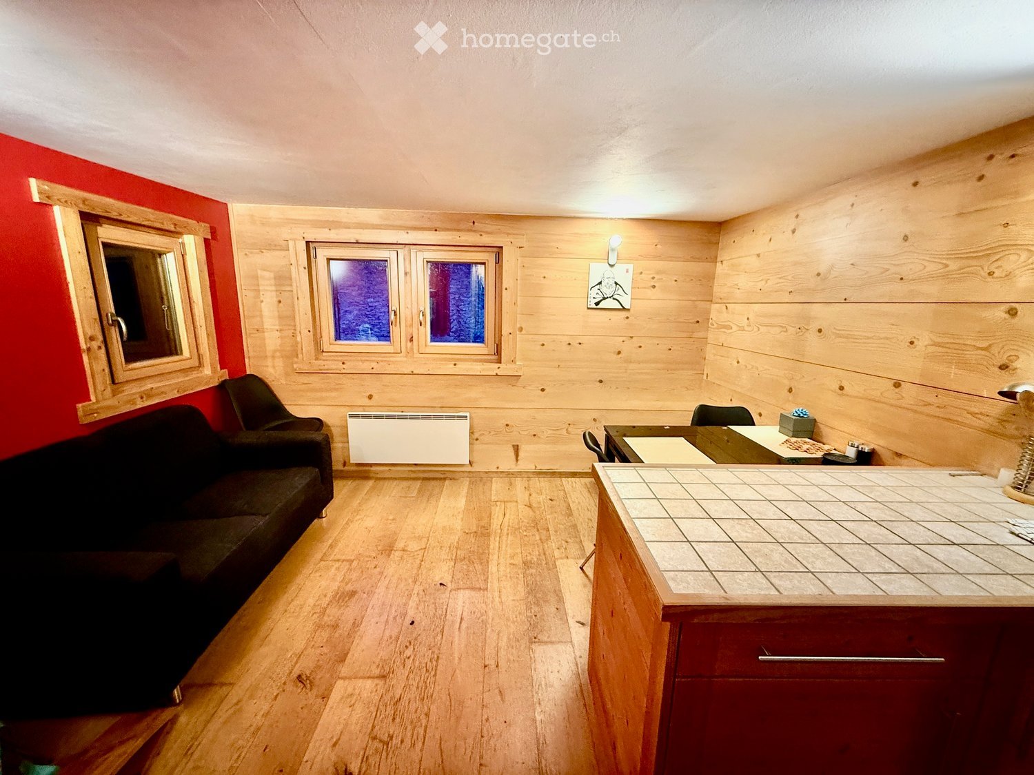 A cozy, wooden-paneled room with a sofa, chair, small table, radiator, mirror, and lamp. The room features wooden flooring and large windows with a view.