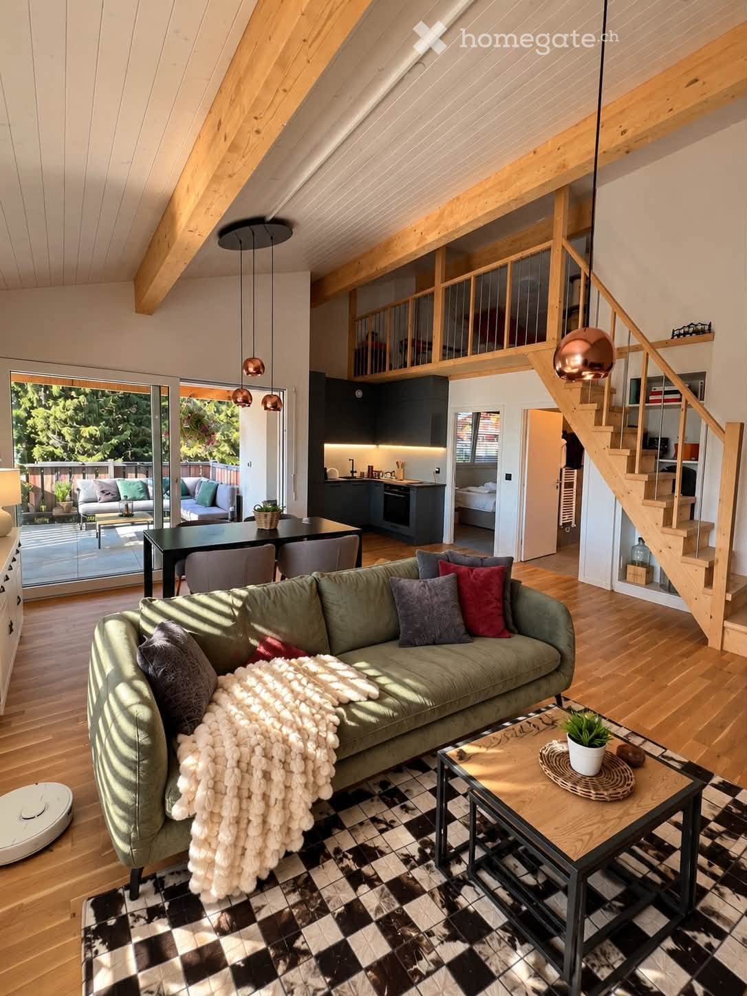 Open-plan living and dining area with a green sofa, coffee table, dining table, and chairs. The space features wooden floors, exposed wooden beams, pendant lights, and a staircase leading to an upper level. A modern kitchen is visible in the background, a