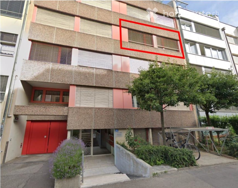 Multi-story apartment building with a red garage entrance, multiple windows, and a small garden area with bicycles and plants.
