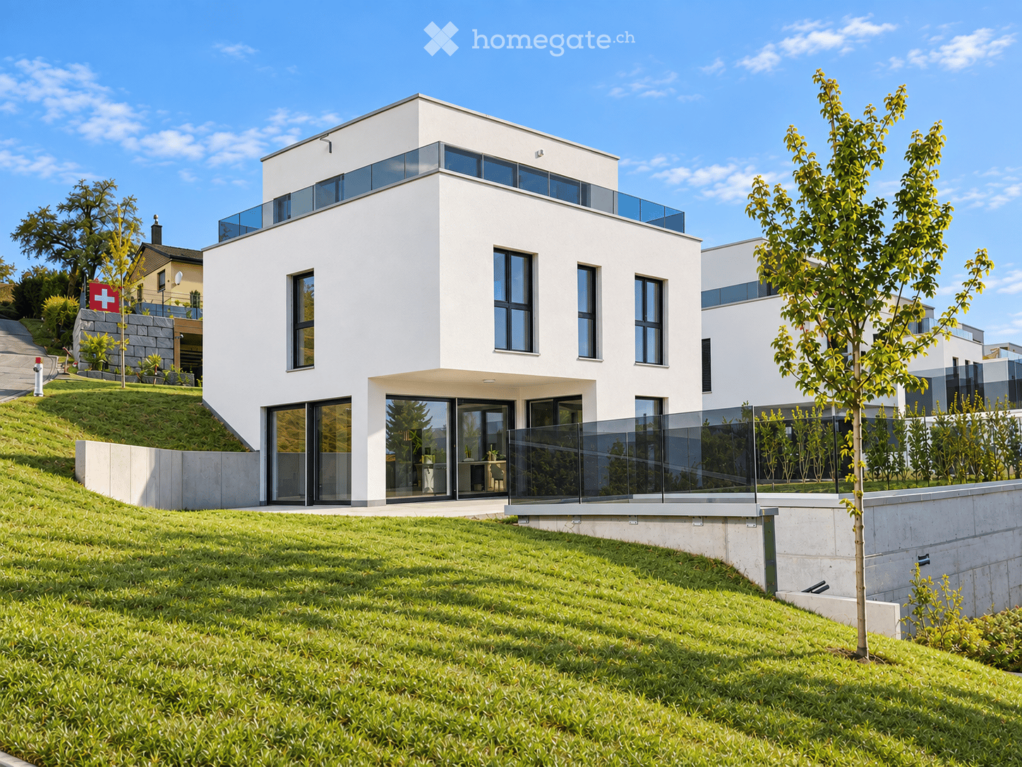 Modern two-story white house with large windows and a raised ground floor, surrounded by a well-maintained lawn and trees.