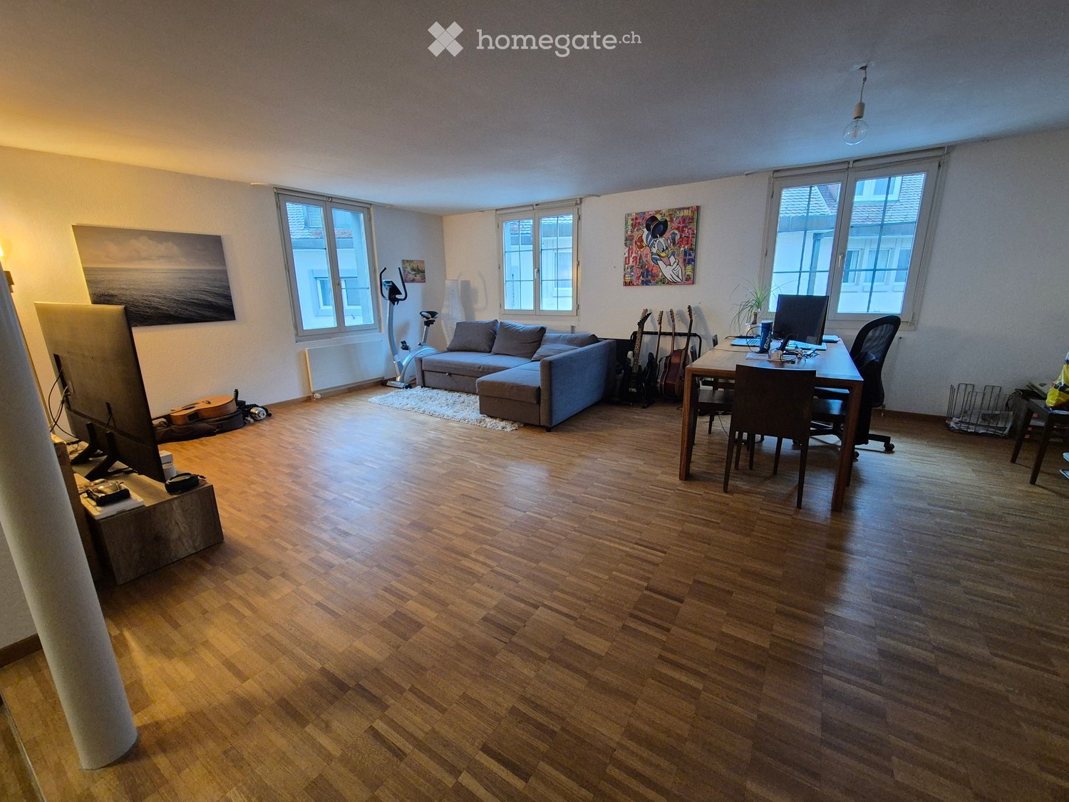 A spacious living room with wooden flooring, large windows, a grey sectional sofa, a dining table with chairs, a TV screen, exercise bike, and guitars.