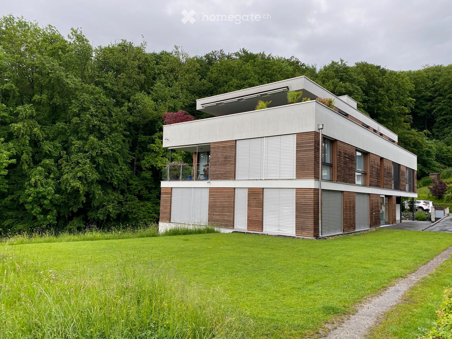 Modern two-story house with white and wood exterior, large windows, balcony, and surrounding greenery.