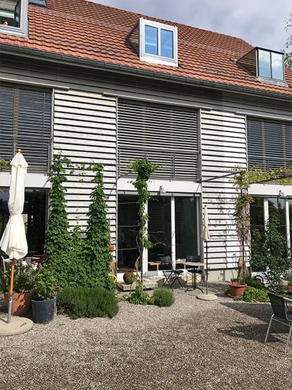 Modern house exterior with a red-tiled roof, white siding, and large windows. The front yard features potted plants, a small patio area with a table and chairs, and a covered outdoor seating area with an umbrella.