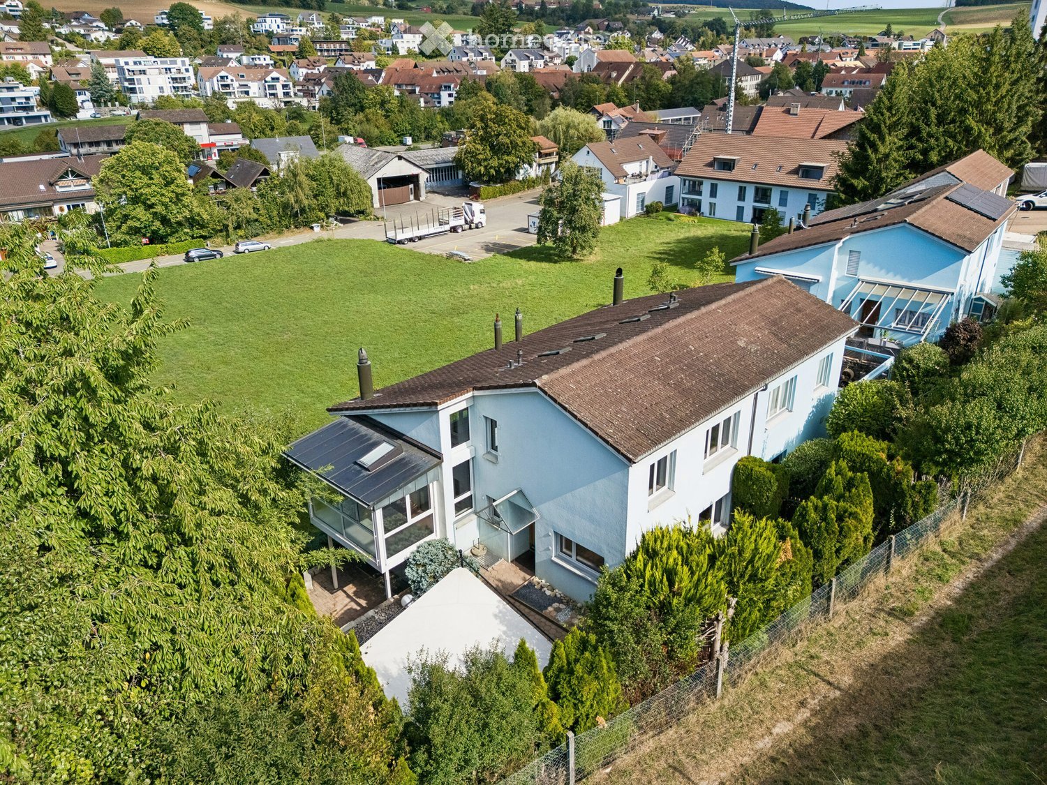 Aerial view of a modern two-story house with a brown tiled roof, surrounded by greenery and neighboring houses in a suburban area.