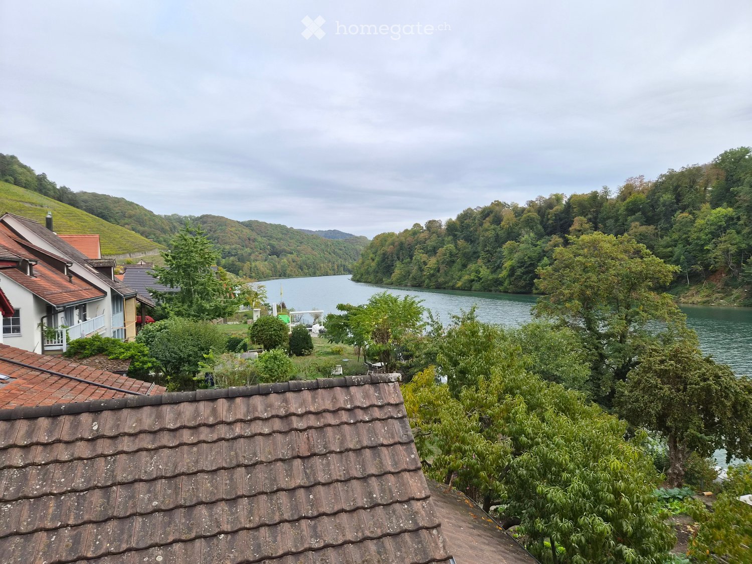 View from a rooftop showing a serene lake surrounded by lush greenery and hills, with a few houses visible on the left side.