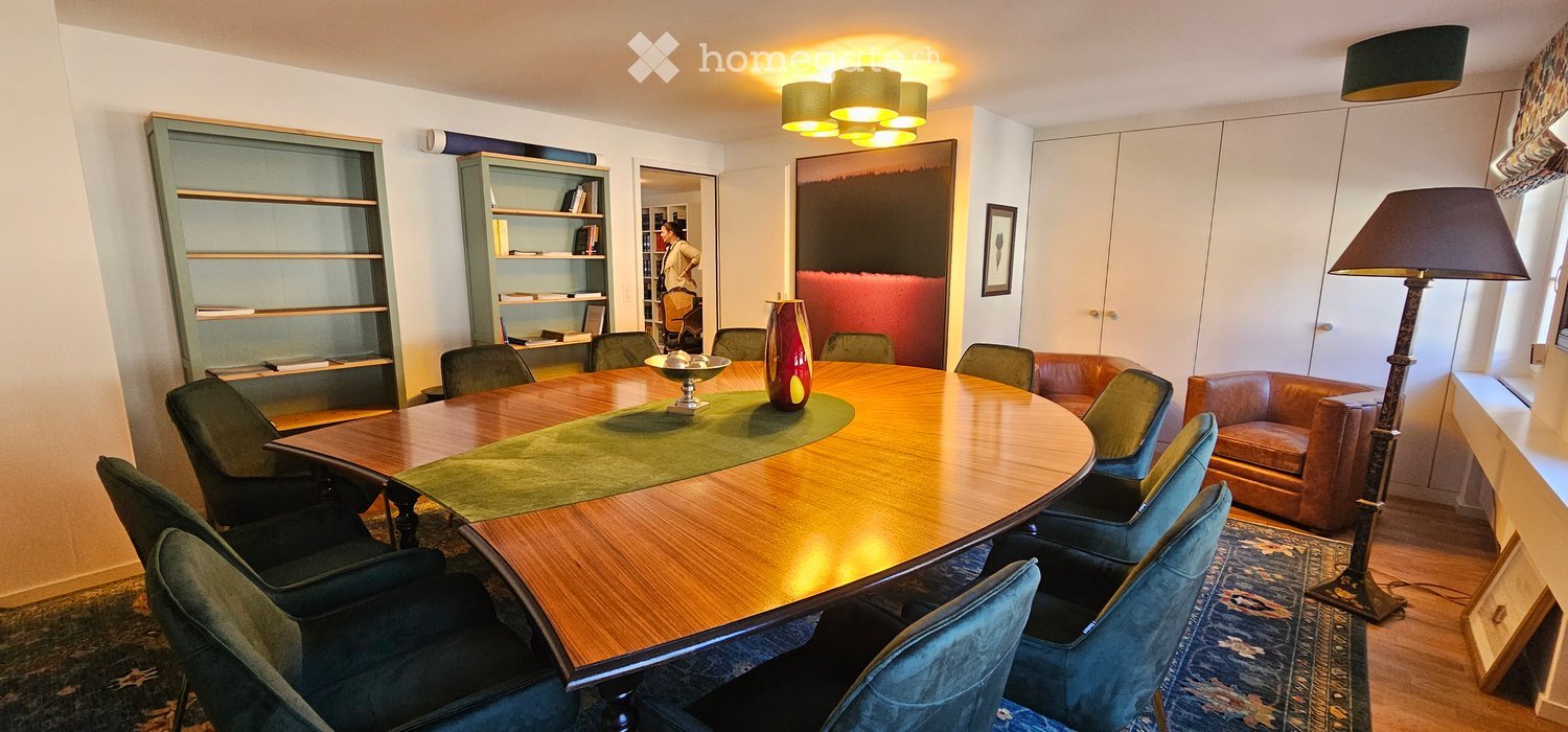 A modern, well-lit room with a large wooden conference table surrounded by green chairs, a leather sofa, bookshelves, and a decorative vase on the table.