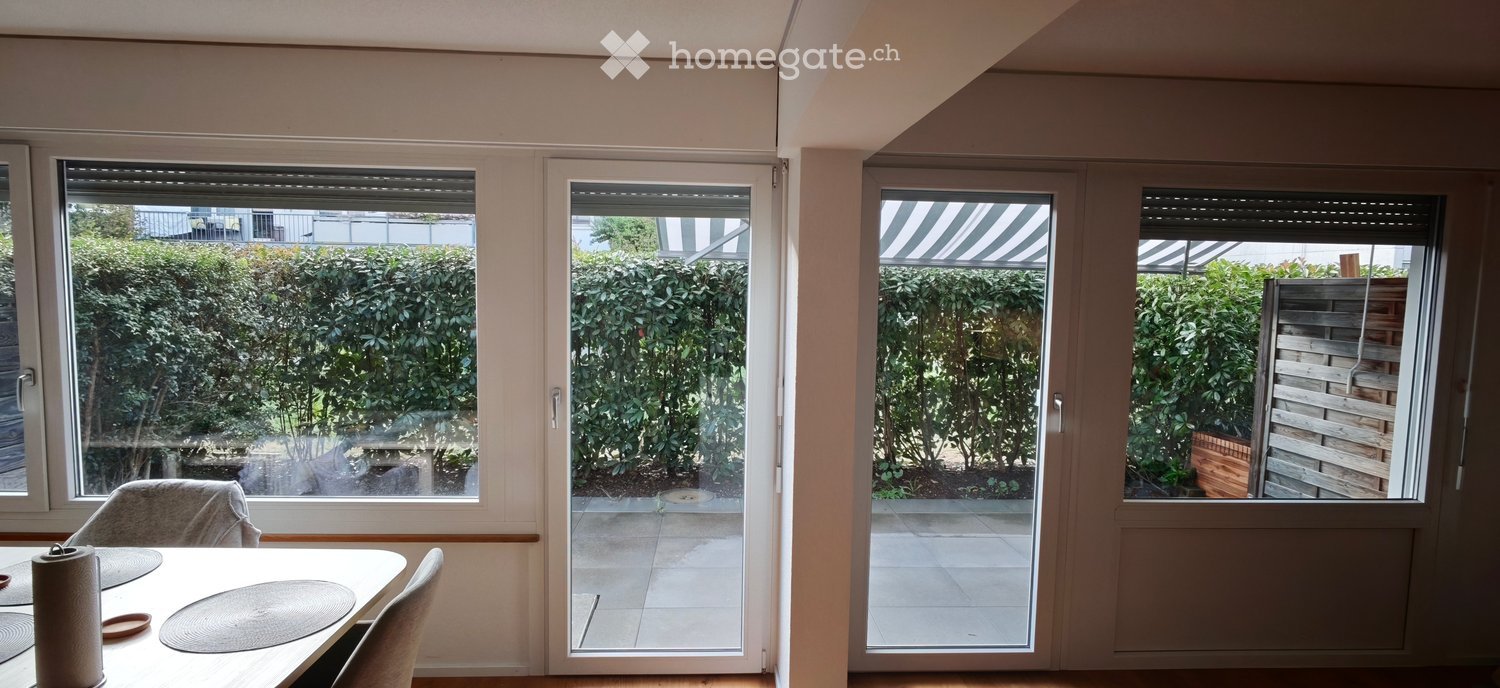Bright dining area with large windows and sliding glass doors leading to a balcony or terrace. The room features a dining table with chairs and a view of a well-maintained garden.