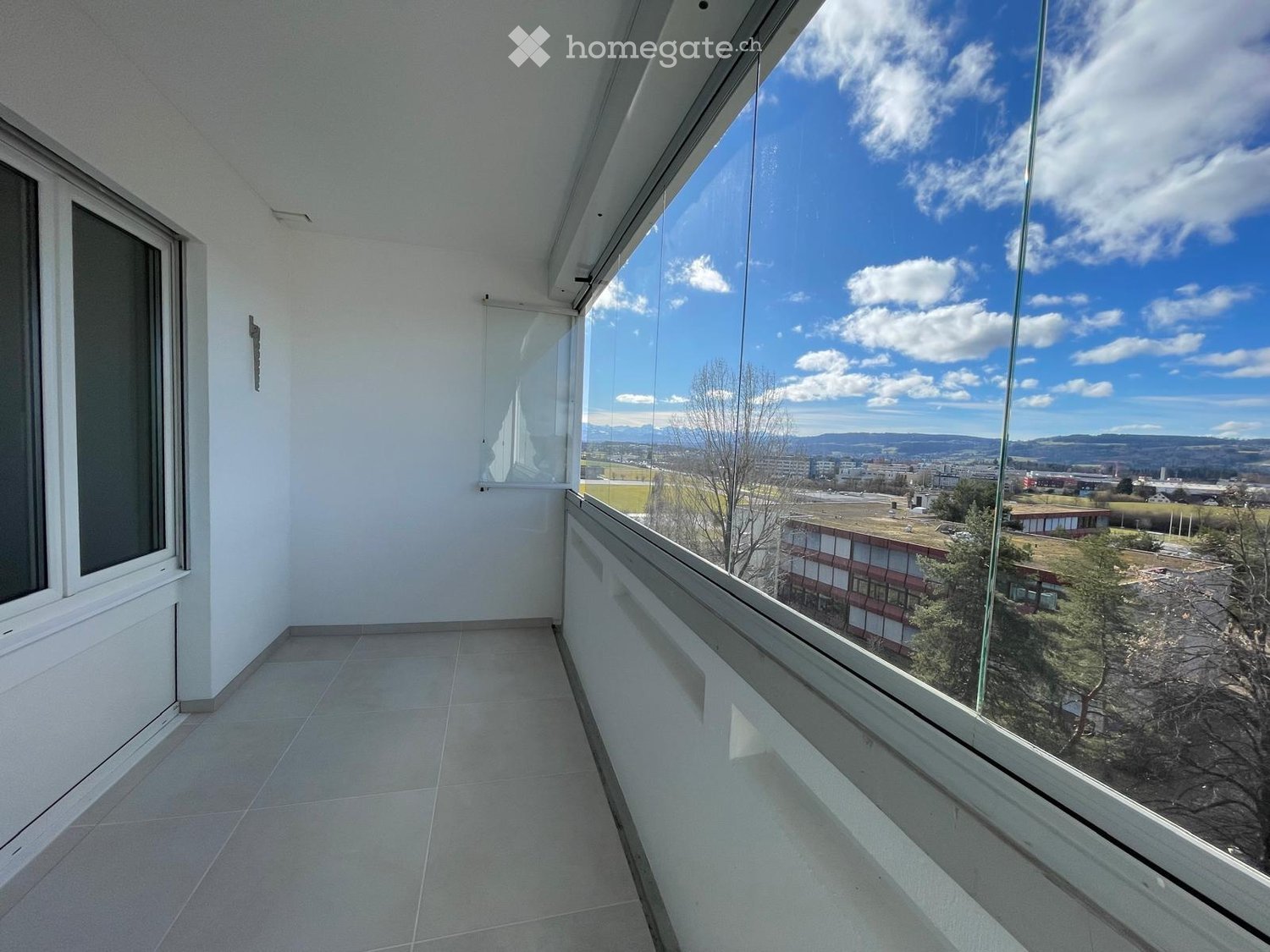 Modern balcony with large glass windows, tiled floor, and a view of the city and surrounding landscape.