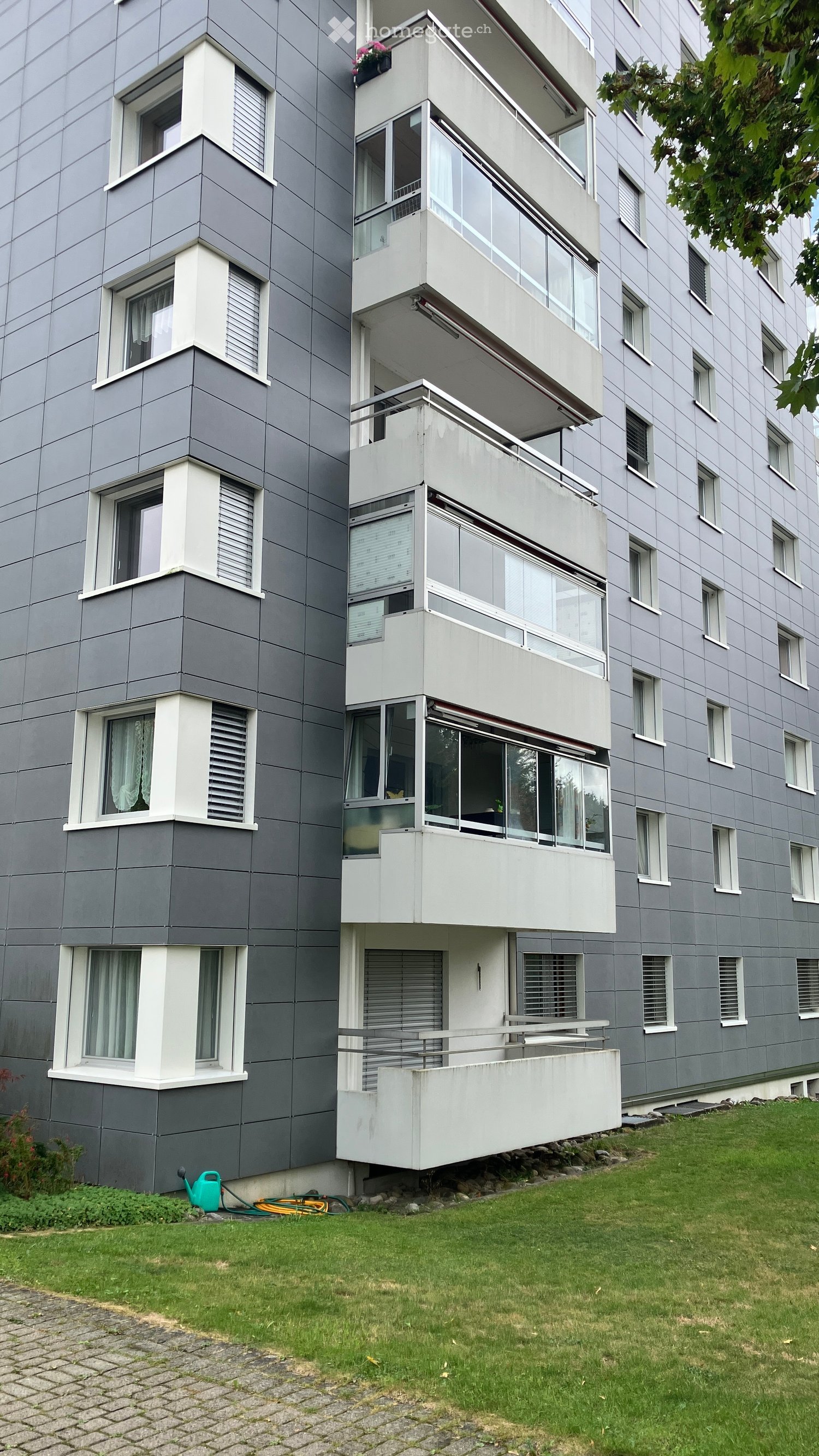 Modern multi-story apartment building with grey and white facade, large windows, and balconies. Green lawn and garden area in front.
